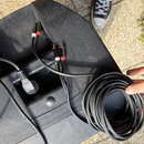 Na černém plastovém povrchu leží srolovaný černý kabel a dva krátké propojovací kabely s červenými a bílými RCA konektory. V prohlubni je vidět černý…
