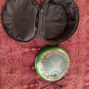 Zelený kovový ruční buben (steel tongue drum) s vryty ve tvaru jazyků na horní ploše. Na boku je viditelný nápis „AAG C D E F G A B C“. Součástí je č…