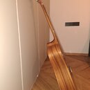 Fotografie zachycuje akustickou kytaru z pohledu ze strany, opřenou o bílou stěnu na dřevěné podlaze. Nástroj má přirozenou dřevěnou barvu s viditným…
