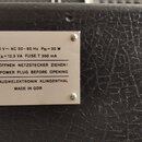 Detail kovového štítku připevněného na černý povrch s texturou kůže. Na štítku je nápis: 220 V ~ AC 50-60 Hz PN = 30 W, PA = 12,5 VA FUSE T 200 mA, V…