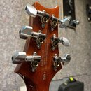Zadní strana hlavy kytary z hnědého lakovaného dřeva s šesti chromovanými ladičkami. Na dřevě je bílý nápis: „built by Cortek Musical Instrument Co.,…