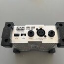 Stříbrná aktivní DI box s černými bočními elementy. Na horním panelu jsou nápisy DAP AUDIO, ADI-100 ACTIVE DIRECT INJECT BOX a ATTENUATION s přepínač…