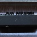 Boční pohled na černý elektronický klavír nebo syntetizátor s dřevěnými bočními panely. Na horní straně jsou vidět řady černých otočných ovladačů a b…