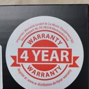 Kruhová bílá nálepka na černém povrchu krabice. Obsahuje červený nápis WARRANTY, ve středu červený pruh s bílým textem 4 YEAR a v dolní části opět ná…