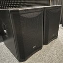Dva černé reproduktory v hranatých bednách s kovovými mřížkami. Na pravém dolním rohu každého reproduktoru je viditelné malé stříbrné logo s nápisem…