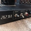 Detailní pohled na černý kovový panel kytarového zesilovače. Na panelu je bílý nápis VK 212 TM a dva přepínače s popisky POWER (ON) a STANDBY (ON). V…
