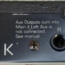 Detail zadní strany černého rackového zařízení s bílými nápisy ENSONIQ a MR-RACK. Jsou zde vidět konektory typu jack označené jako R Aux, L Mono, R a…