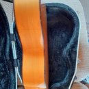 Fotografie zachycuje zadní stranu akustické kytary v hnědém lakovaném provedení s viditnou strukturou dřeva. Nástroj leží v otevřeném tvrdém kufru s…