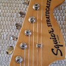 Fotografie zachicuje hlavu elektrické kytary z přírodního dřeva s chromovanými ladičkami. Na hlavě je černý nápis Squier STRATOCASTER® a pod ním menš…