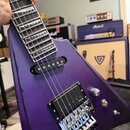 Fialová elektrická kytara s hranatým tělem a hrazdicími pražci. Na obou snímačích je bílý nápis EMG a na kobylce je viditelný nápis Floyd Rose. Povrc…