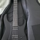 Černá elektrická kytara s matným povrchem v černém softcasu. Nástroj má dva humbuckery, fixační most a na hlavě není vidět žádné logo ani nápis. Povr…
