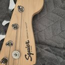 Záběr na hlavu basové kytary z přírodního světlého dřeva. Na zadní straně jsou čitelné nápisy Squier BY FENDER a BRONCO BASS®. Jsou zde vidět čtyři k…