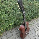 Akustická kytara v tmavě hnědé barvě s matným povrchem. Nástroj stojí na černém stojanu před zeleným živým plotem na dlažbici. Na hlavě kytary nejsou…