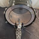 Černý basový buben s chromovaným obručováním a bílým průhledným koženem. Na bubnu je nápis REMO POWERSTROKE3 BASS a zlatý štítek s nečitelným nápisem…
