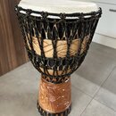 Djembe s bílou membránou a dřevěným korpusem. Povrch dřeva je zdoben řezbami a černými ornamentálními vzory. Nástroj je opleten černou šňůrkou s bare…