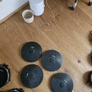 Čtyři černé gumové pady elektronických činelů s koncentrickými drážkami, položené na dřevěné podlaze. Na každém padu je bílý nápis ALESIS. V levé čás…