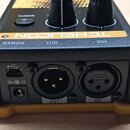 Zadní strana černého elektronického zařízení s žlutým základním rámem. Jsou zde vidět konektory USB, napájecí vstup a dva XLR konektory s popisky OUT…