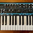 Černý analogový syntetizátor s klaviaturou a ovládacím panelem. Na levé straně je nápis novation BASS STATION II Analog Synthesizer. Panel obsahuje m…