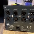 Tmavý kovový box s audio konektory. Na horní části jsou čtyři výstupy označené OUTPUT 4, OUTPUT 3, OUTPUT 2 a OUTPUT 1 a čtyři vstupy označené INPUT…