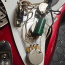 Fotografie zachycuje vnitřní elektroniku elektrické kytary s červeným korpusem a bílým plastovým krytem. Jsou vidět zapájované potenciometry, zelený…
