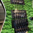 Detail elektrické kytary s zeleným plamenovitým povrchem. Nástroj má dva černé humbuckery s bílými nápisy „Jackson“ a černý tremolo systém s nápisem…
