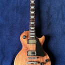 Elektrická kytara s tónem dřeva v přirozené barvě a černým pickguardem. Na hlavě je viditelné logo Gibson. Nástroj má dva humbuckery, čtyři ovládací…