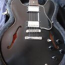 Černá polodupatá elektrická kytara s černým pickguardem a dvěma humbuckery. Nástroj je umístěn v otevřeném kufru s šedým polstrováním. Na těle jsou č…
