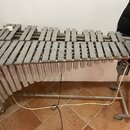 Stříbrný vibrafon s kovovými pleskami a trubkami rezonátory umístěný na kovovém stojanu s kolečky. Nástroj je vybaven elektrickým kabelem a bílým ová…