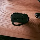 Černý elektronický přístroj s obaleným černým suchým popruhem. Na horní straně jsou vidět USB-C port a kulatý konektor pro sluchátka. Nad USB portem…