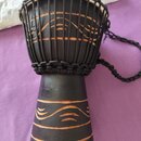 Černý djembe s bílou membránou, nataženou pomocí černých provázků. Tělo nástroje je zdobeno vyrytými vodorovnými a vlnitými liniemi, které odhalují h…