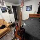 Elektrická basová kytara s hnědým průsvitným lakem a tmavě červeným pickguardem. Na hlavě nástroje je svisle napsáno logo "Music Man". Nástroj má čty…