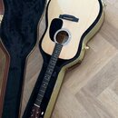 Akustická kytara s přírodním dřevem a černým plektrem v hnědém tvrdém kufru. Na hlavě nástroje je viditelné logo "Martin". Povrch nástroje i kufru vy…