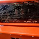 Zadní strana kytarového komba v oranžovém vinylu s černou kovovou mřížkou, pod kterou jsou vidět elektronické válvy. Na spodním černém panelu jsou ná…