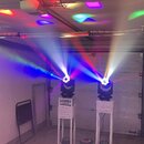 Dvě černé svítící hlavy umístěné na bílých kovových stojanech. Světla vyzařují barevné paprsky v modré, červené a bílé barvě. Na levém spodním zaříze…