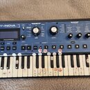 Modrý synthesizer s klaviaturou a nápisem MININOVA SYNTHESIZER a logem novation. Přístroj obsahuje displej, řadu tlačítek, otočných ovladačů a osm čí…