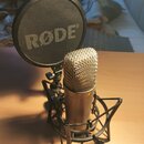 Kondenzátorový mikrofon v kovovém provedení s mřížkovou hlavicí, umístěný v černém pavoukovém držáku. Před mikrofonem je umístěn kulatý černý popfilt…