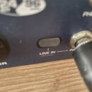 Detailní záběr na modré kovové šasi audio zařízení. Je vidět vstup s nápisem LINE IN (REAR) a část nápisu PHONES v horní pravé části. V levém horním…