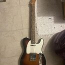 Elektrická kytara typu Telecaster s tónem sunburst (přechod od černé k hnědé a žluté). Má bílý pickguard, dva snímače a na hlavě hmatníku je viditeln…