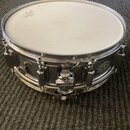 Stříbrný kovový snare drum s bílou blánou. Na bláně je viditelné logo znázornující korunu a nápis REMO. Povrch kovového těla vykazuje známky opotřebe…