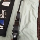 Fotografie zachycuje tmavě hnědou elektrickou kytaru z profilu, vedle které leží černý obal s bílým nápisem Gibson USA. Na kytaru jsou vidět čtyři ov…
