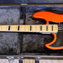 Oranžová elektrická basová kytara v černém tvrdém kufru. Na hlavě hmatníku je nápis Marcus Miller. Nástroj má černý pickguard, dva snímače a světle h…