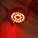 Černý kulatý LED reflektor s integrovanou rukojetí, který svítí červeným světlem. Zařízení obsahuje několiku kruhově rozmístěných LED diod a je připo…