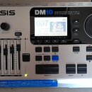 Stříbrný elektronický modul k bubnům s nápisem ALESIS a označením modelu DM10. Zařízení obsahuje displej, několik ovládacích tlačítek a čtyři posuvní…