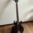 Tmavě červená elektrická basová kytara s černým plechem a černou hlavou. Na hlavě je zlaté logo Gibson. Nástroj stojí v černém stojanu na dřevěné pod…