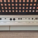 Zadní strana klavíru v béžové barvě s černou mřížkou nahoře. Jsou zde vidět různé konektory a popisky: LCD CONTRAST, TO HOST, MIDI HOST SELECT, IN MI…