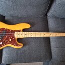 Žlutá elektrická basová kytara s hnědo-červeným tortieshell pickguardem. Na hlavě nástroje je nápis Fender PRECISION BASS. Kytara má čtyři struny a s…