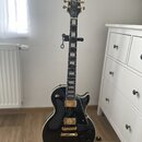 Černá elektrická kytara s gold hardwarem a bílým bindingem, umístěná na stojanu. Na hlavě hmatníku je bílý nápis Epiphone. Kytara má dva humbuckery,…