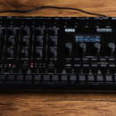 Černý hybridní bicí automat KORG drumlogue. Zařízení obsahuje množství otočných knoflíků, několik tlačítek a malý displej s textem BASS DRUM. Na horn…