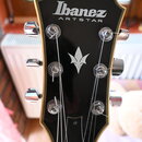 Čelní pohled na hlavu kytary v černé barvě s bílým lemováním. Na čele je bílý nápis Ibanez a pod ním ARTSTAR, v dolní části je bílý symbol stylizovan…