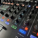Detailní pohled na černý DJ mixér s mnoha ovládacími knoflíky a tlačítky. Na panelu jsou viditelné nápisy jako CUE, FILTER, AUX, CUE VOL, CUE MIX a T…